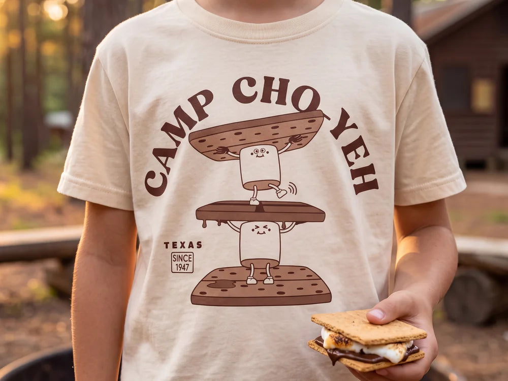 A camper at the fire ring wearing the cream CAMP CHO-YEH s’more tee and holding a s’more