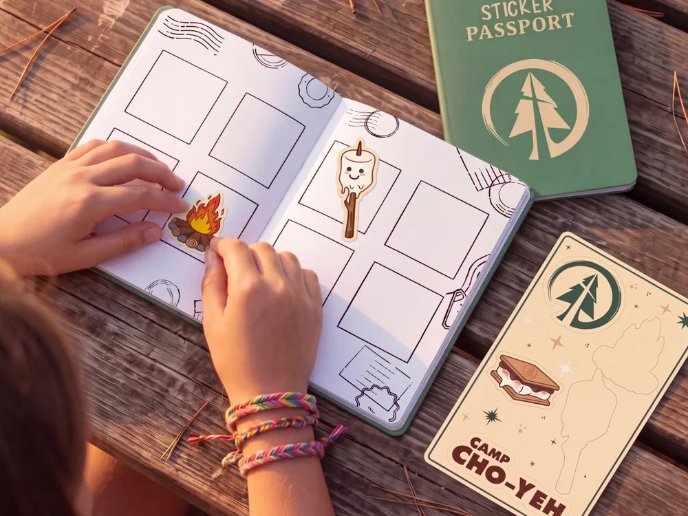 A camper at a picnic table pressing stickers into the open Sticker Passport, with the green cover and sticker sheet beside it